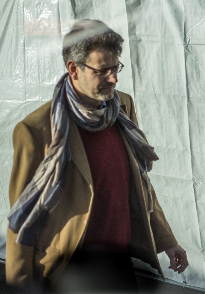 Seen through a window, the scarf, the hand, the face in shadow, I wonder who he is.